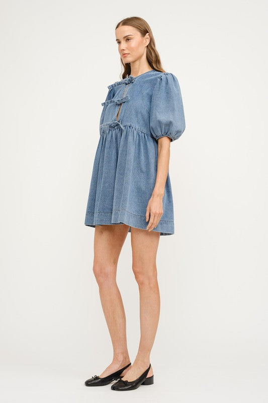 A denim dress in a medium denim wash, featuring three front ties at the chest, a loose fit with seem at the waist creating a gentle waist line and a gentle puff sleeve. A perfect dress for every day wear this spring, graduation parties, and casual nights out.