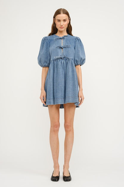 A denim dress in a medium denim wash, featuring three front ties at the chest, a loose fit with seem at the waist creating a gentle waist line and a gentle puff sleeve. A perfect dress for every day wear this spring, graduation parties, and casual nights out.