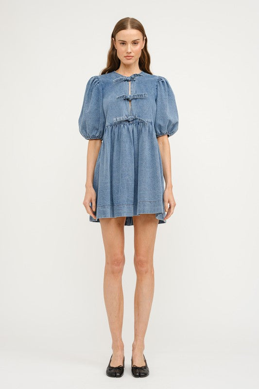 A denim dress in a medium denim wash, featuring three front ties at the chest, a loose fit with seem at the waist creating a gentle waist line and a gentle puff sleeve. A perfect dress for every day wear this spring, graduation parties, and casual nights out.