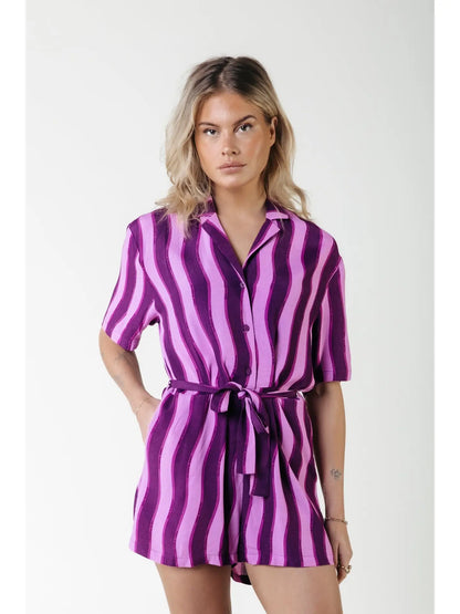 Bright purple romper featuring bold dark purple and light purple stripes with a waist wrap to tie in the front, button up romper.